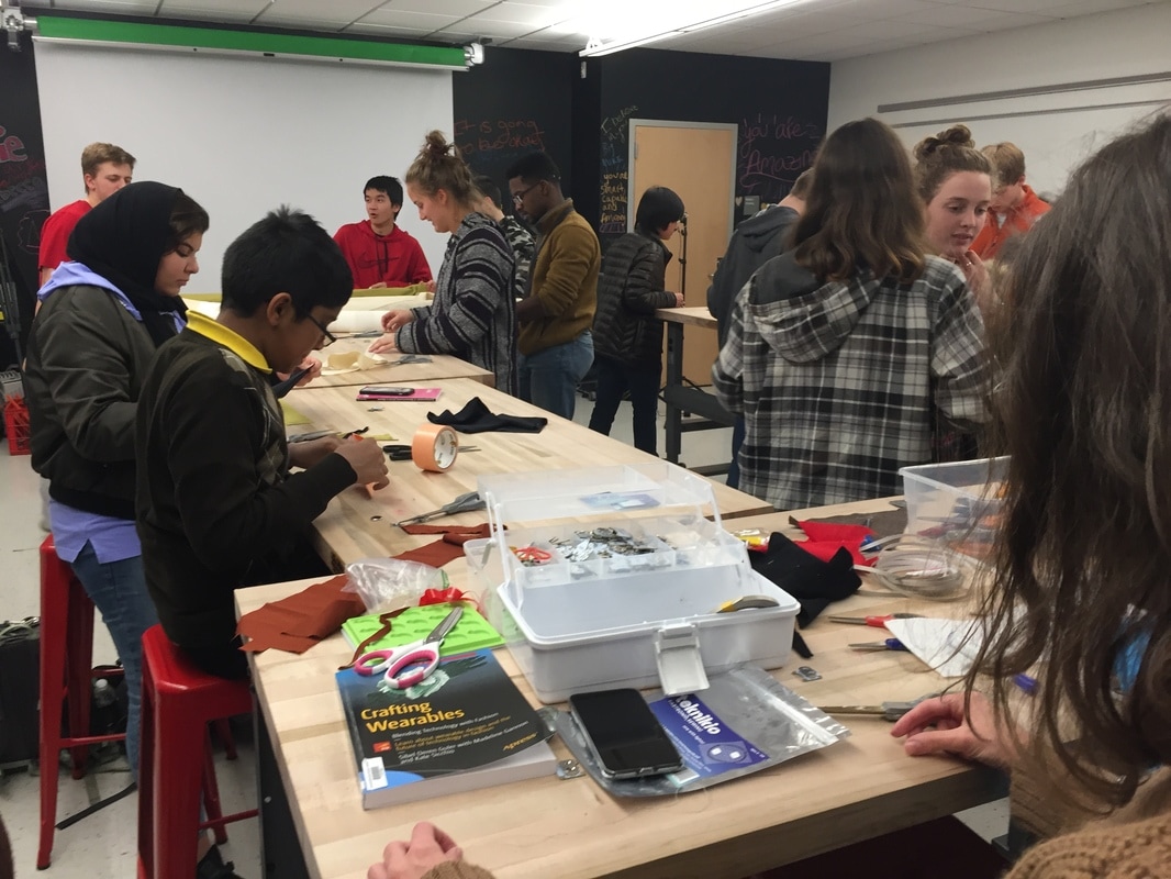 A group of students work at tables to create wearable tech. Some students are seated, others are standing. There are containers and supplies all over the tables, and a book on how to create wearable tech.