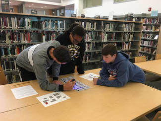 Three students (one female and two males) are clustered around a worksheet on a table. They are using a UV pen to try and solve the puzzle.