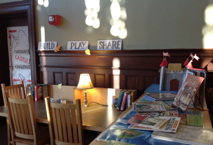 There is a table with books in the foreground, and a table with chairs and a lamp in the background. There are also words displayed on the wall that read 