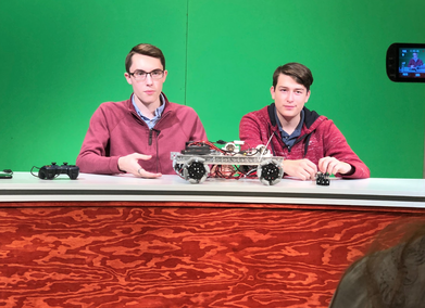 Two male presenting students wearing red jackets are showing off a robotic creation on a table. They are in front of a green screen. There is a camera in the corner that is recording them.