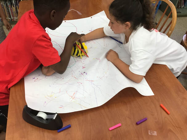 A boy and girl are leaning on a table that has a piece of white paper, tape and crayons.  They are both examining a yellow and black plastic toy on top of the paper.