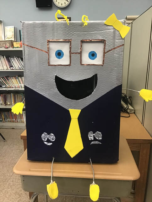 CalBOT is a robot made out of cardboard and has a black colored bottom and gray top.  CalBOT has square framed gold glasses, a yellow tie and yellow hands and feet.