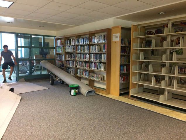 Library space with a rolled gray carpet on top of a gray carpet.  Two library cases one full with books and one empty.  A student helper in the doorway.