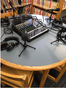 A sound mixing board sits on a table surrounded by multiple microphones and headphones. They are on a round table. There are book cases in the background.