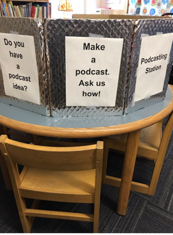 Three signs on shiny silver paper are set up trifold style on a table. The middle sign reads make a podcast ask us how. The left sign reads do you have a question? And the right hand sign reads podcast station.