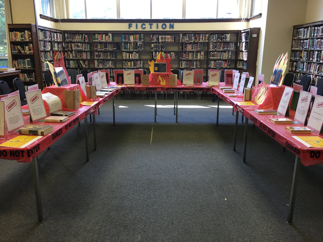 Three tables are set up in a U shape - there are signs explaining why each book was banned behind each book on the table, as well as tiered displays with fake flames.