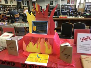 Books are arranged on a table wrapped in brown paper with a caution stamp across the front. There is also a tiered display that looks like flames popping out behind an explanation of the banned books display.