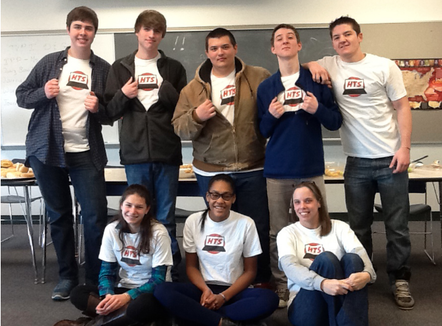 A group of seven high school students, five boys and two girls, pose with their teacher. The students are part of the teen tech squad. They are showing off their custom t-shirts. The boys are standing and the girls are on the floor with their teacher.