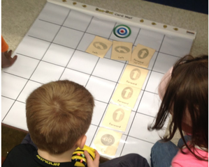 Two students bend over a chart with squares drawn on it. There are yellow cards with directional arrows laid out on the grid and one student holds a Bee Bot robot in their hands. They are attempting to direct the robot to the target.