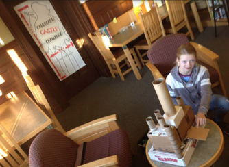 A student is posing with a completed cardboard castle on a table in the library. 