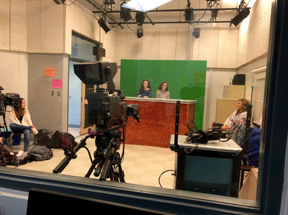 Two students are behind a glass wall in the recording studio. They are in front of a green screen and seated at a desk. Two other students are at the sides of the image. There are mounted lights on the ceiling and a camera in front of the students.