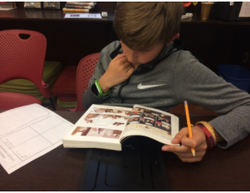 A male-presenting student is sitting at a table. He has a pencil in one hand and is looking at a graphic novel. There is a worksheet next to the book. The student appears deep in thought.