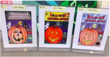 Three coloring pages that have been completed are displayed in plastic stands. The image is of a jack o'lantern with the words 